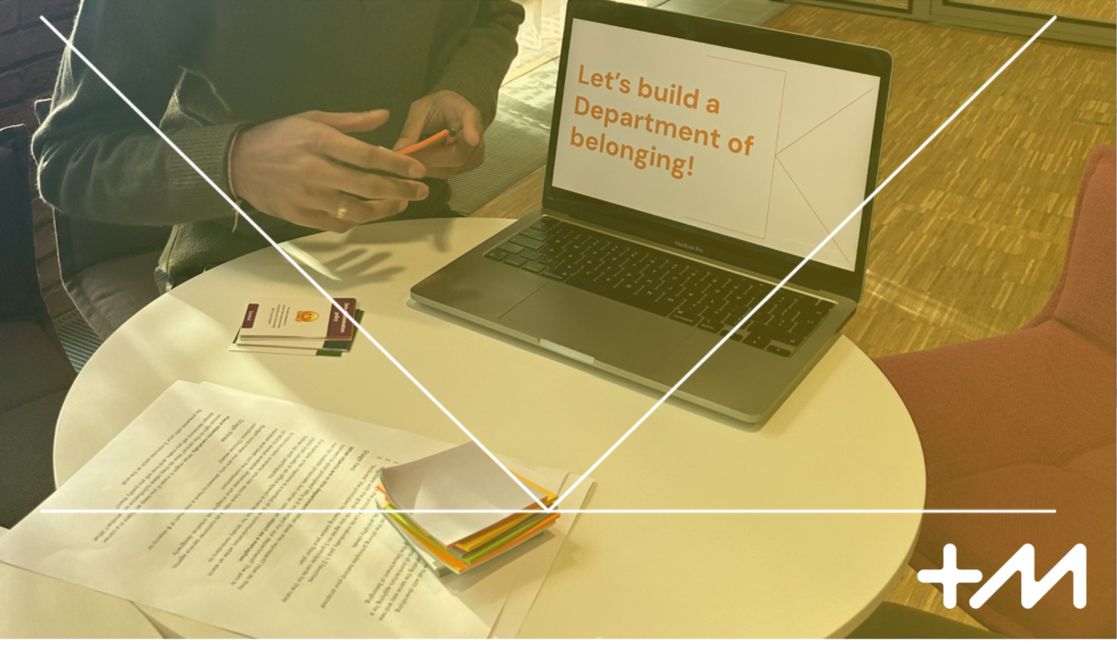 Round small table with some papers, cards and sticky notes piled up, along with a laptop with a slide "Department of belonging" open. There is a person behind the desk, only hands visible.